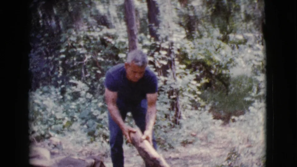 A man in jeans grips a log or branch in a wooded clearing during the 1968 Cottonwood, Arizona home movie.