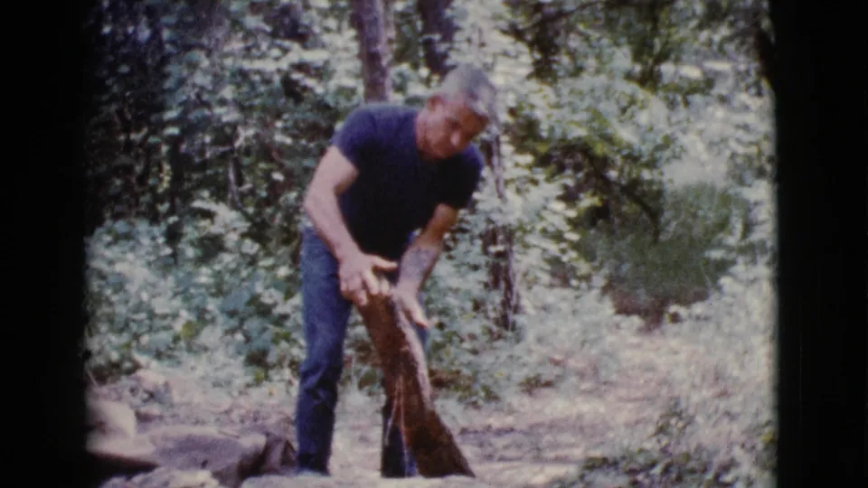 The man raises or steadies a long piece of wood in a wooded Cottonwood, Arizona scene from 1968 8mm footage.