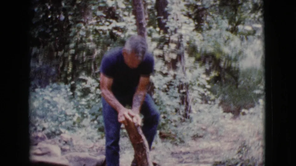 The man continues working the partially cut log in a wooded Cottonwood, Arizona setting, with dense greenery behind him.