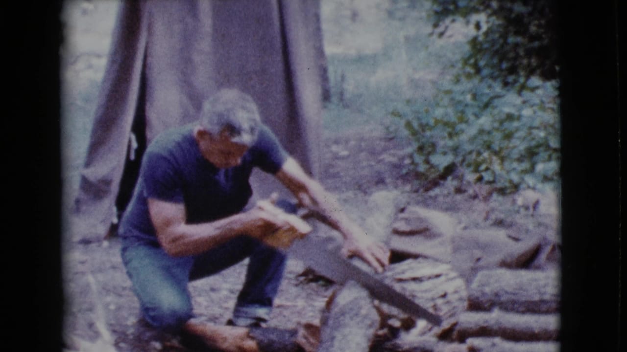 Man crouches beside a log and works a hand saw near a canvas tent.