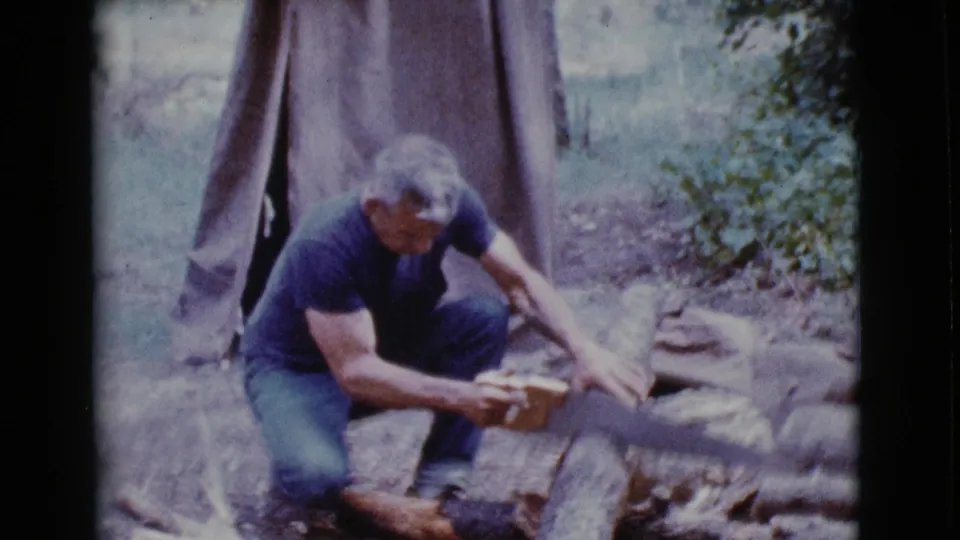 The man continues sawing a log near a canvas tent in the 1968 Cottonwood, Arizona campsite scene.