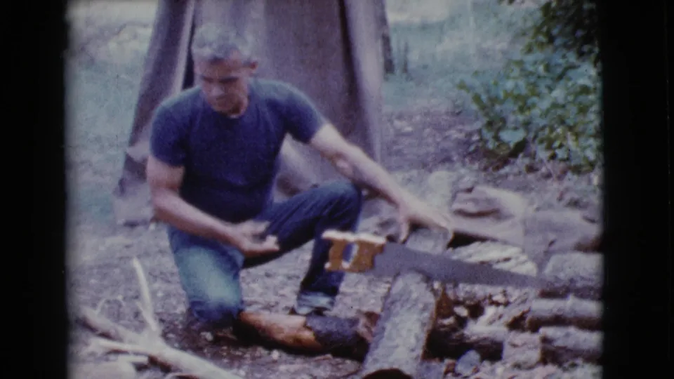 A crouching man holds a hand saw across a log, with rocks, brush, and canvas tent fabric visible behind him.