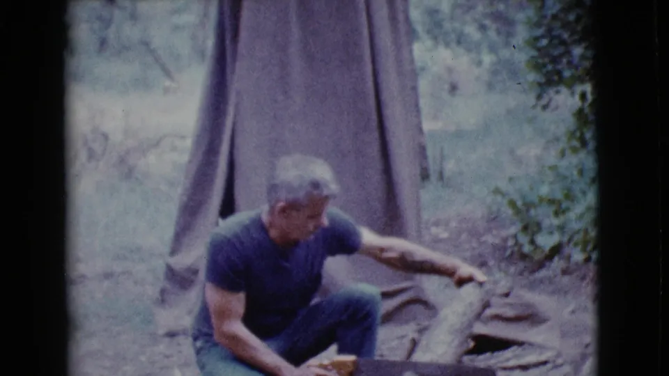 At the end of the clip, the man crouches beside the log near the tent, with the wooded campsite softly visible around him.