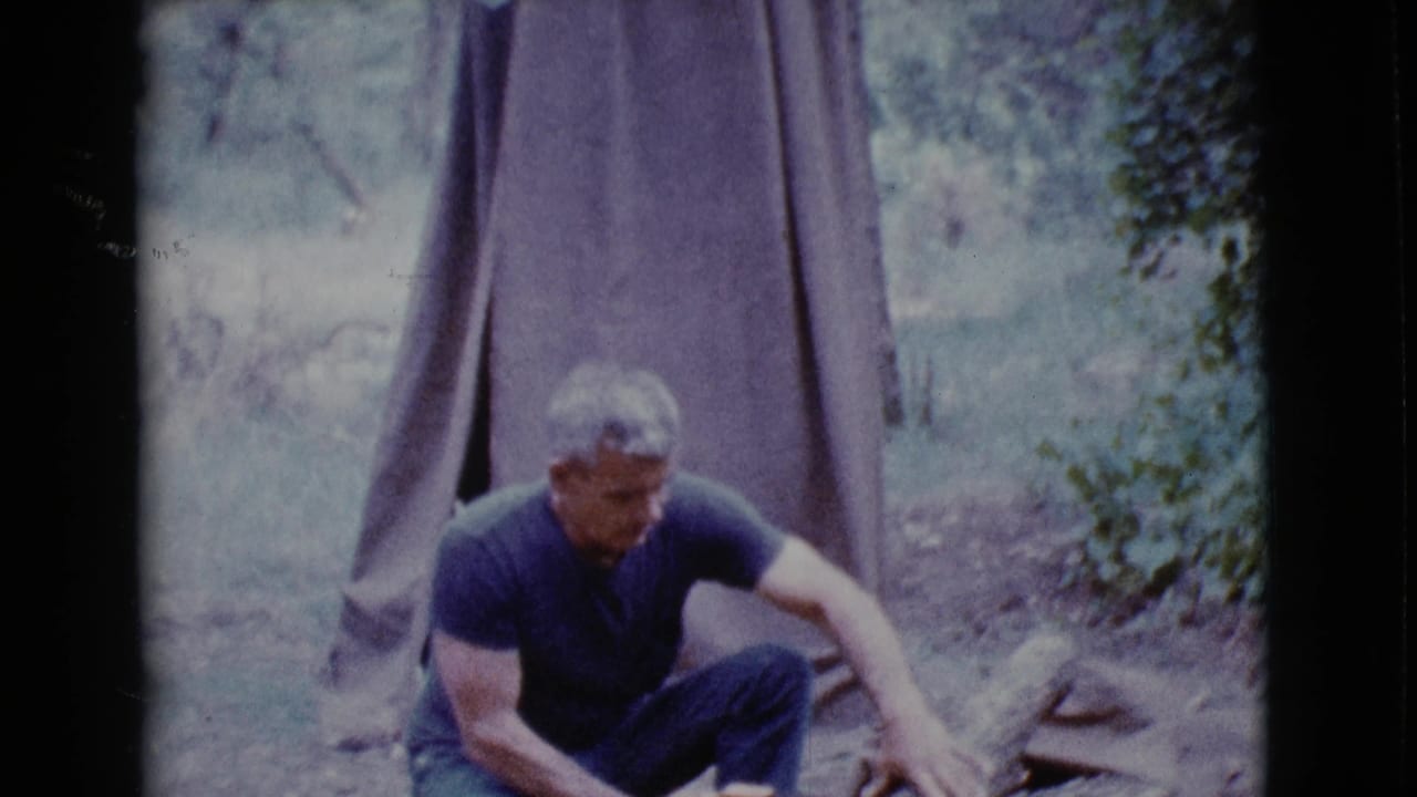Man crouching at a campsite beside a canvas tent and log.