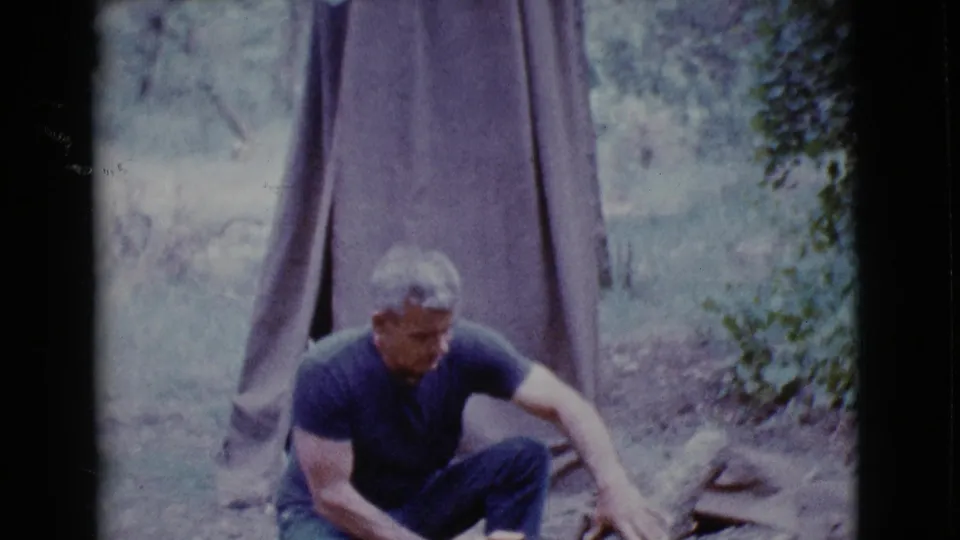 In Cottonwood, Arizona, 1968 footage, an adult man crouches near a log in front of a canvas tent at a campsite.