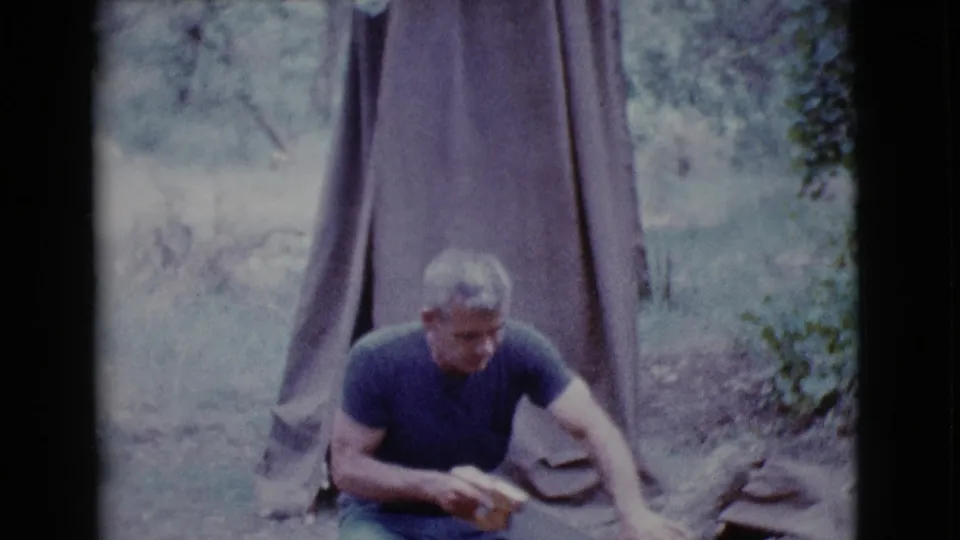 The man holds a hand saw while crouched by a log, with a canvas tent and trees visible in the 1968 Arizona campsite scene.