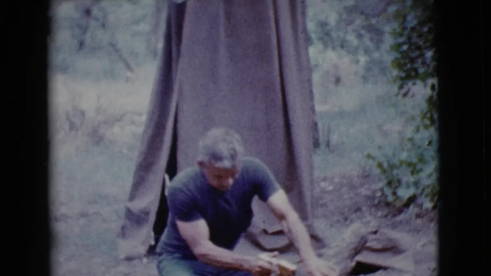 A man leans over the log with a hand saw at a Cottonwood campsite, filmed in soft, faded 8mm color.