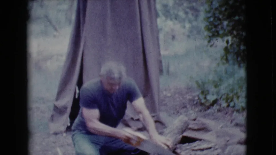 At the Arizona campsite, the man saws a log by hand in front of a canvas tent, with trees and rocks around the camp area.