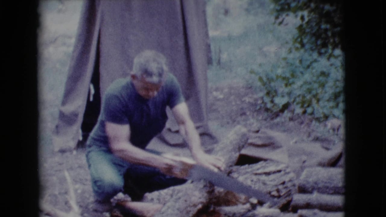 Man kneels beside a log and uses a hand saw near a canvas tent.