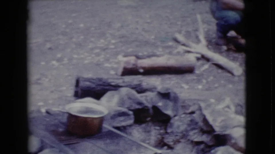 The campsite fire area includes a copper-colored pot, stones, logs, branches, and scattered gear on bare ground.