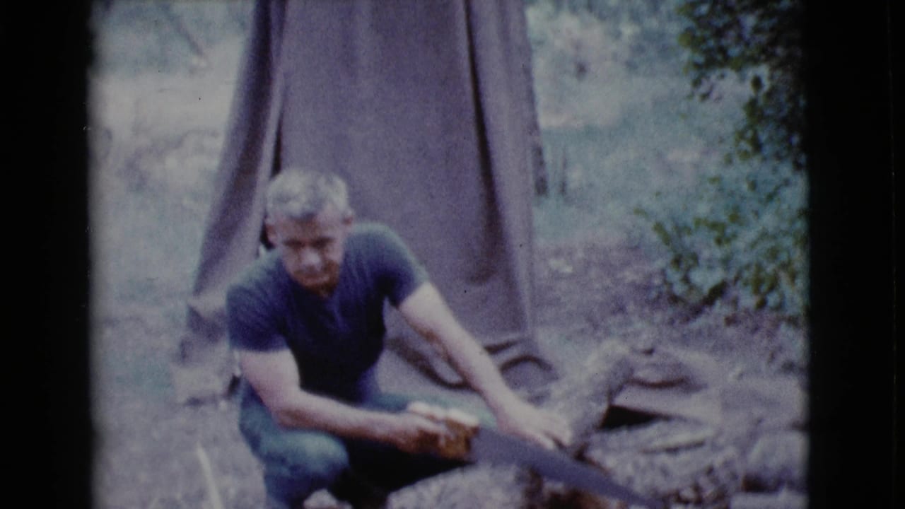 Man crouching beside a log near a brown tent in a wooded area.