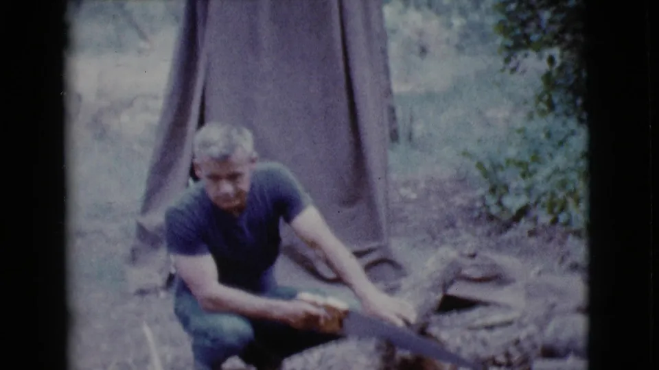In Cottonwood, Arizona in 1968, a man in a blue shirt crouches near a tent while sawing or handling a log.