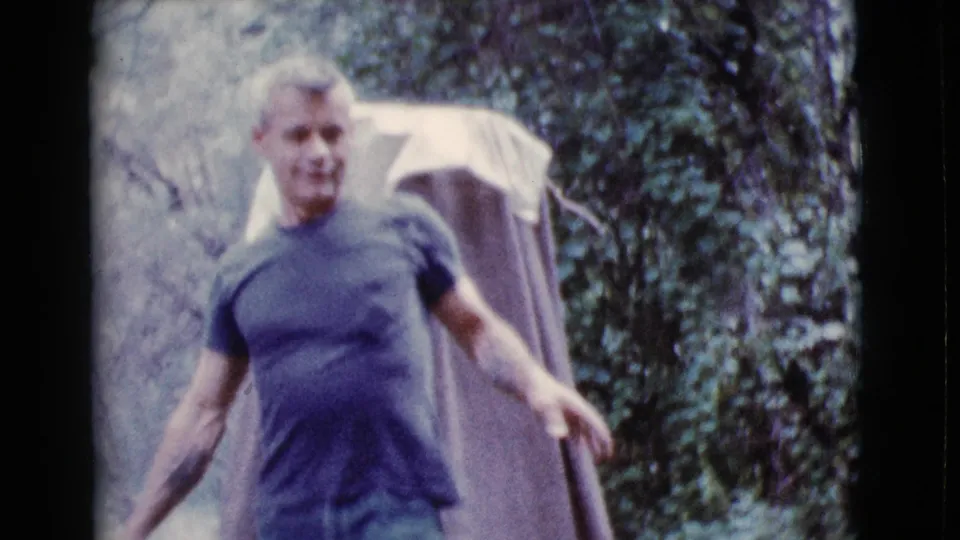 The man smiles toward the camera while moving through the wooded campsite near the tent in Cottonwood, Arizona.