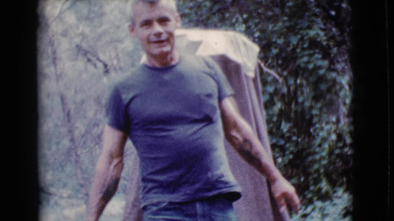 Adult man in a dark T-shirt walks through trees carrying a light folded tent or tarp.