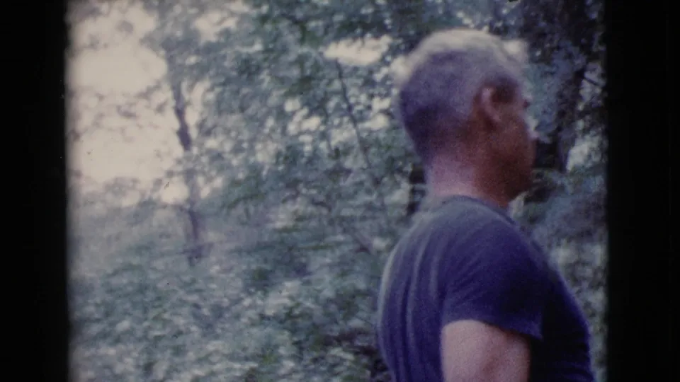 Rear view of an adult man in a dark T-shirt walking away through a wooded Cottonwood, Arizona setting in 1968 home-movie footage.