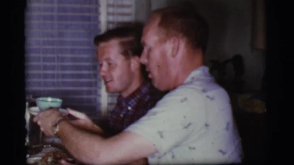 Two men in a 1968 Cottonwood, Arizona home kitchen sit at a table near window blinds, with cups and food in front of them.