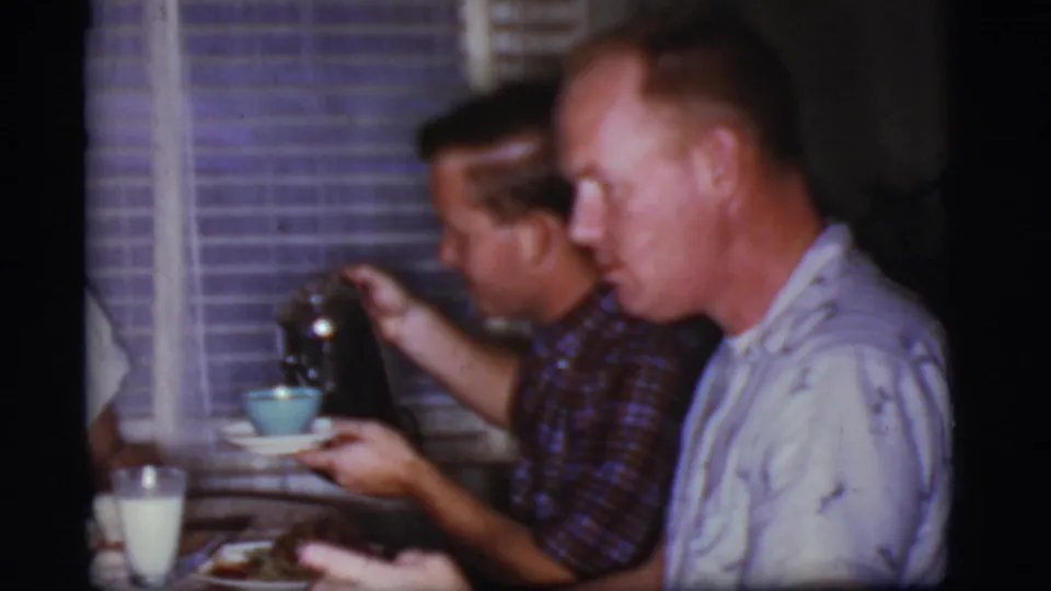 In the 1968 kitchen scene, a seated man pours tea from a dark pot into a light blue cup while another man eats nearby.