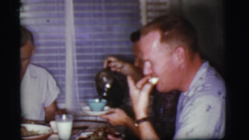 A man drinks from a small cup in the Cottonwood kitchen while another seated man holds the dark teapot near the table setting.
