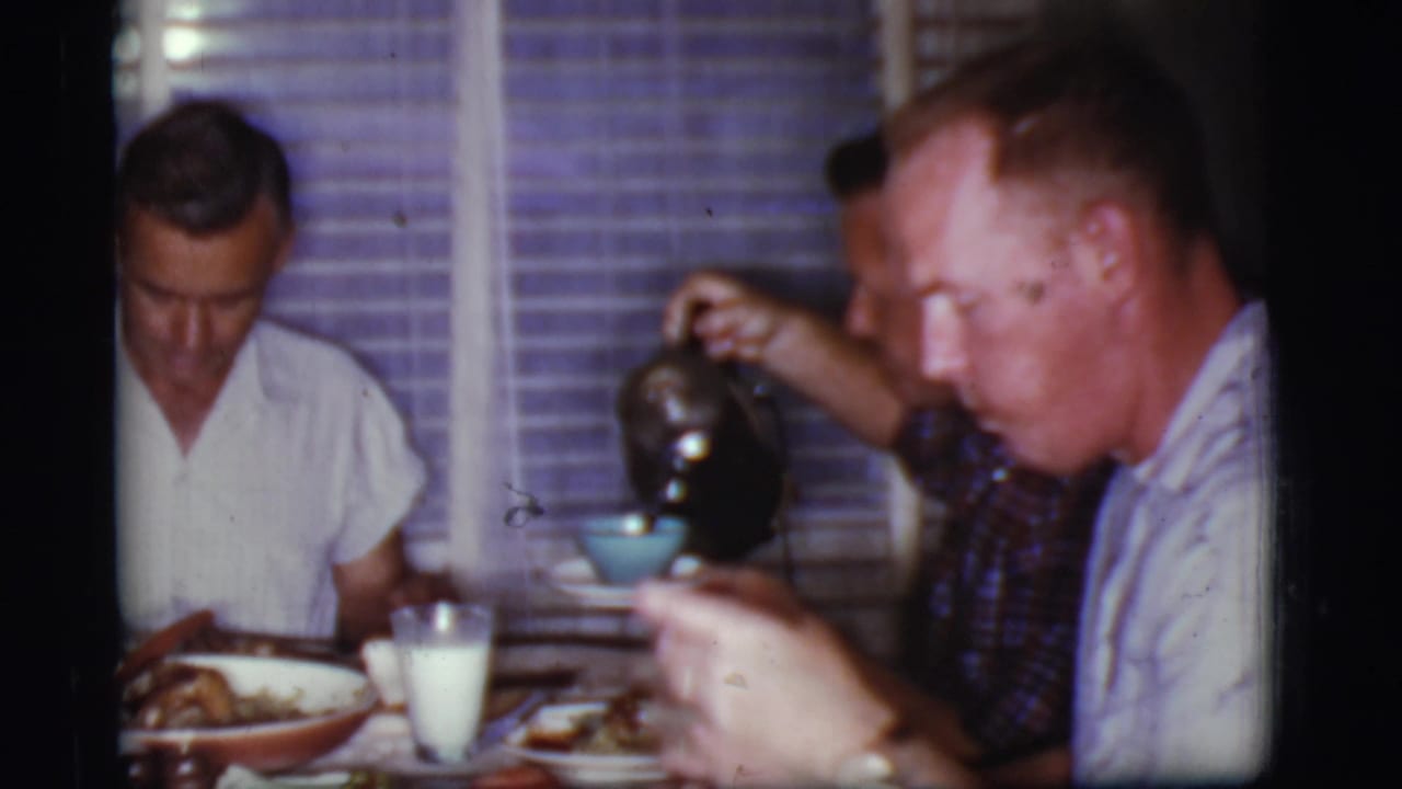 Three men seated at a dinner table as one holds a dark coffee pot near a blue cup.