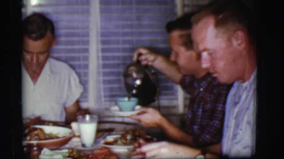 1968 home-movie frame from Cottonwood, Arizona showing a man pouring coffee into a blue cup beside two men eating indoors.