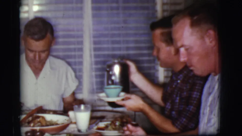 Three adult men sit at an indoor dinner table in 1968 Cottonwood, Arizona footage as coffee is held over a blue cup.