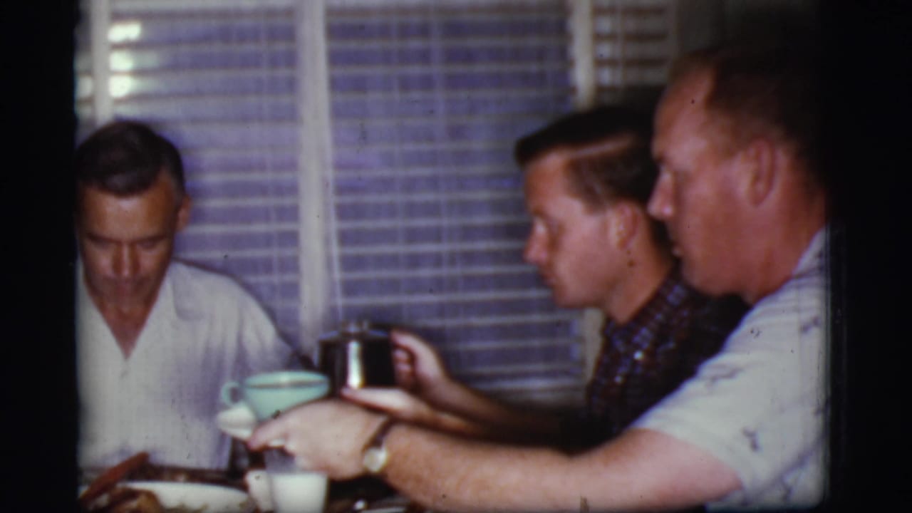 Three adult men sit at a breakfast table while one passes a cup near a metal coffee pot.
