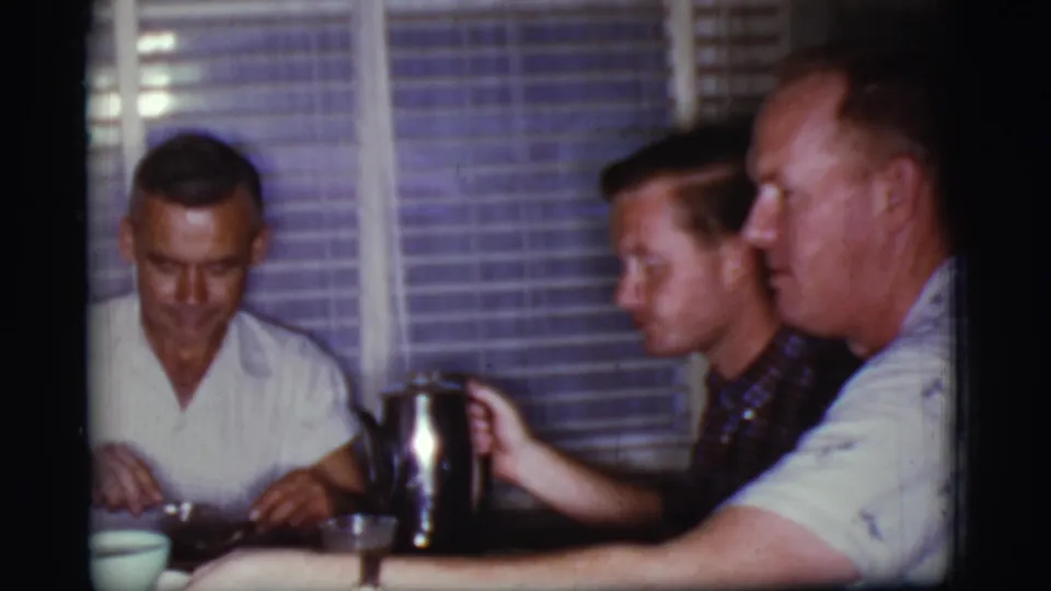 1968 Cottonwood, Arizona frame showing men seated at breakfast, with a hand near a metal coffee pot and window blinds behind them.