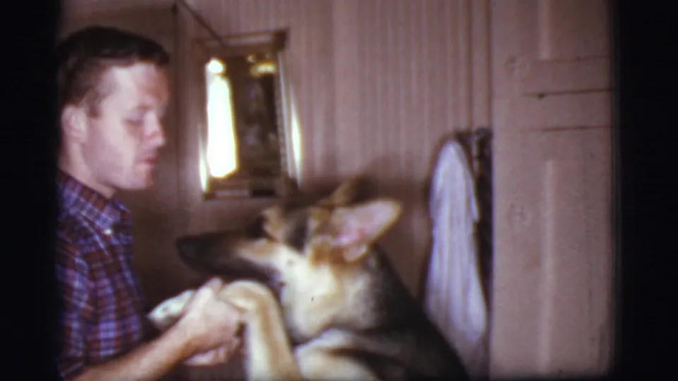 A man in a plaid shirt faces a German Shepherd with raised paws in a Cottonwood, Arizona private interior, filmed on 8mm in 1968.