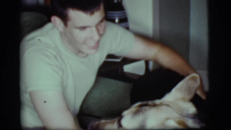 A smiling man in a light T-shirt pets the German Shepherd at close range inside the 1968 Cottonwood, Arizona home.