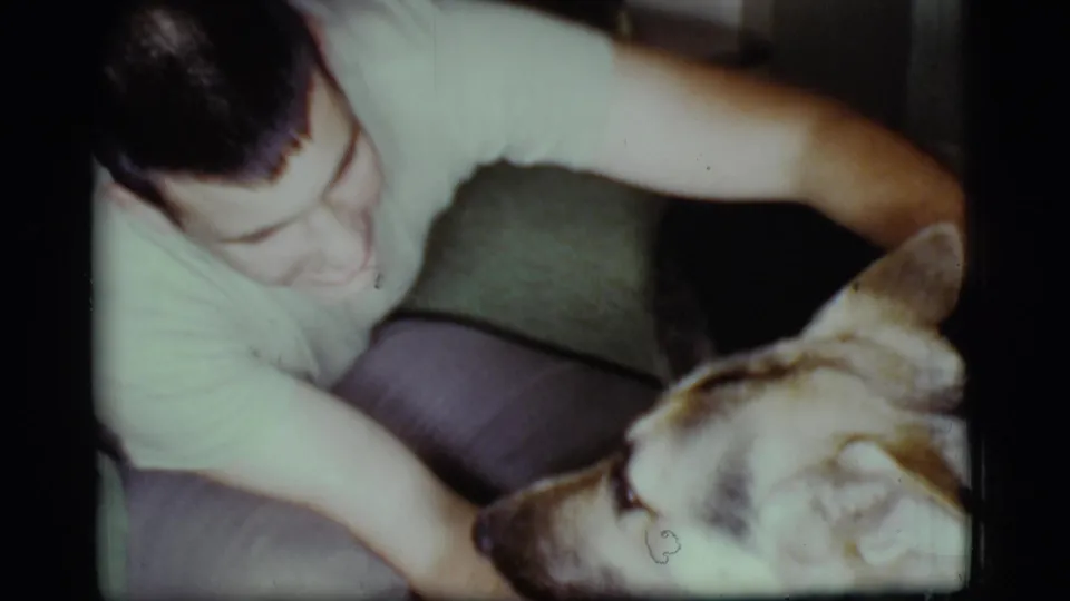 In a 1968 Cottonwood, Arizona home interior, a young man smiles while petting a German Shepherd resting on a couch.