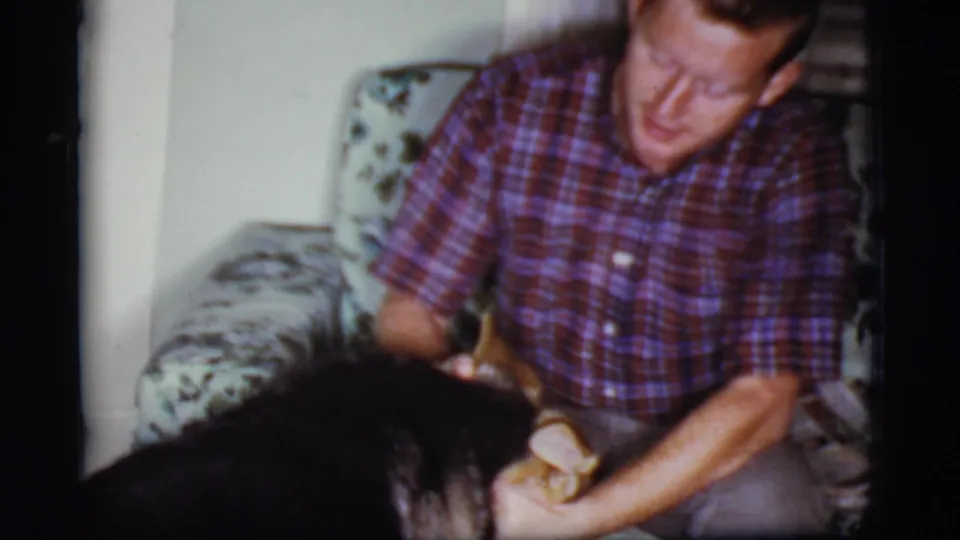 In 1968 Cottonwood, Arizona home-movie footage, a seated man in a plaid shirt pets a German Shepherd on a floral sofa.