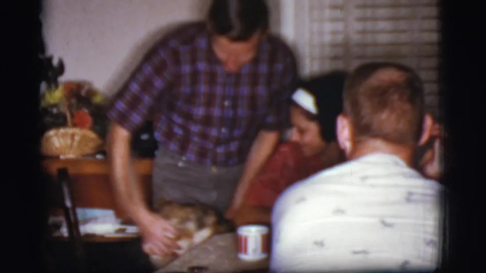 The plaid-shirted man bends toward the German Shepherd beside a table, with seated adults visible in the private interior.