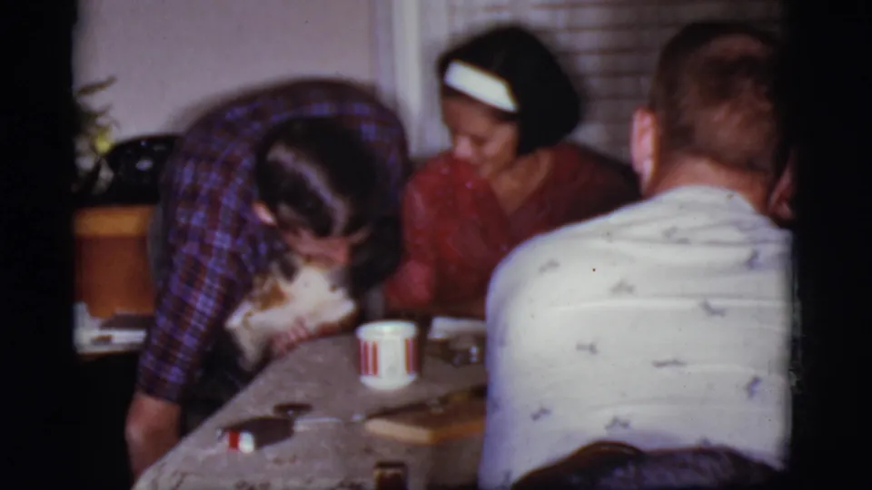 A 1968 home-movie frame shows adults at a table while the man bends to pet or guide the German Shepherd indoors.