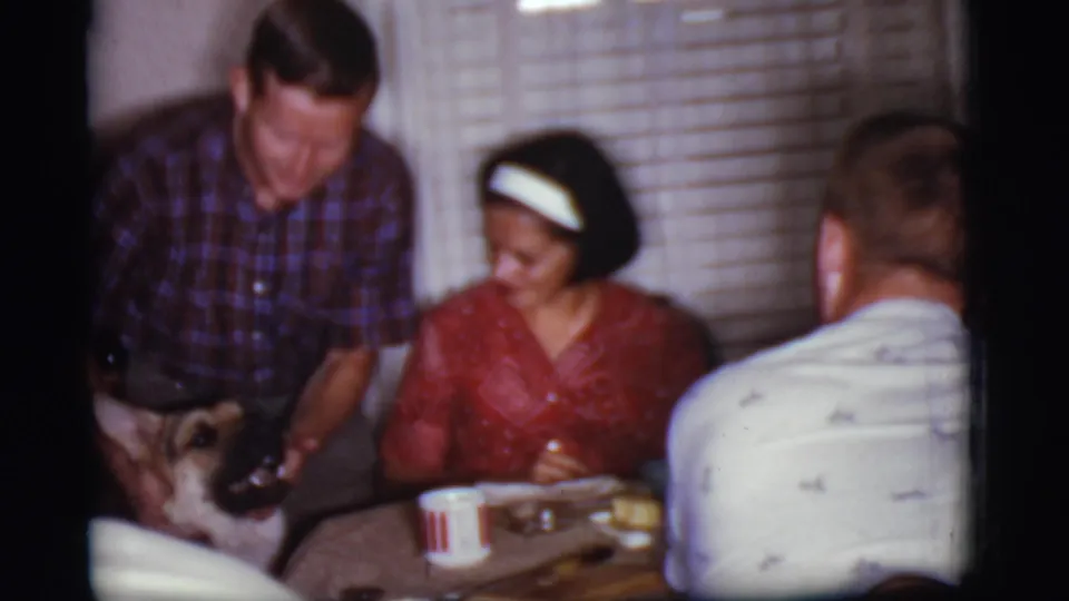1968 Cottonwood, Arizona frame showing a man petting a small dog near a dinner table with adults seated nearby.