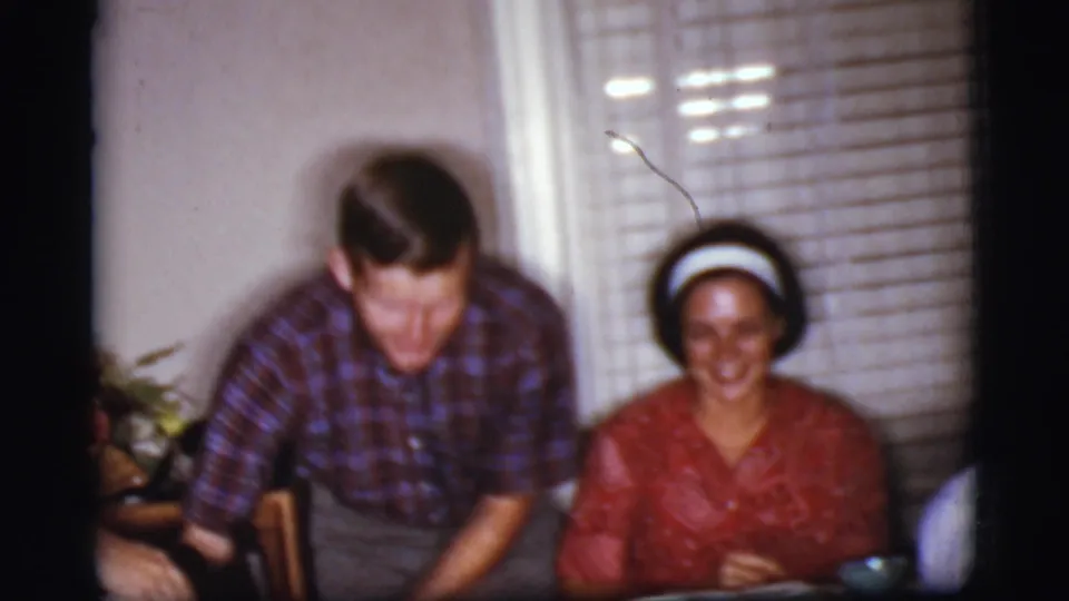 1968 Cottonwood, Arizona home-movie frame of a smiling woman in red seated at a table as a man stands nearby.