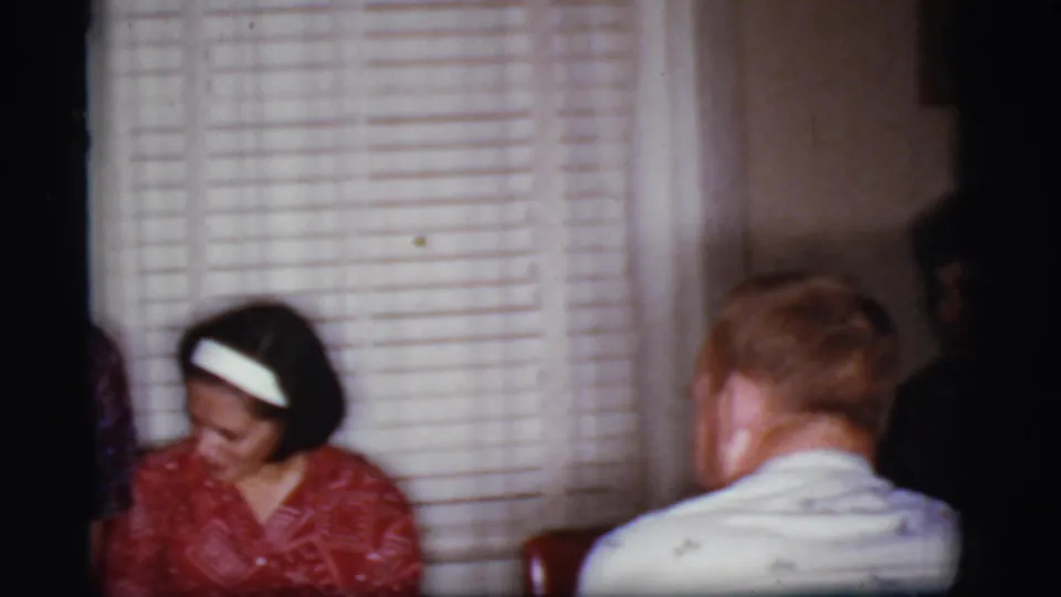 1968 Cottonwood, Arizona frame of a woman in a red dress seated near slatted blinds, facing a man seen from behind.