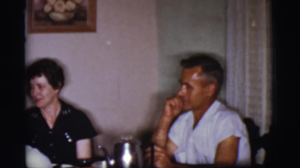 1968 Cottonwood, Arizona frame of a woman in a dark dress and a man in a white shirt seated at a dining table inside a home.