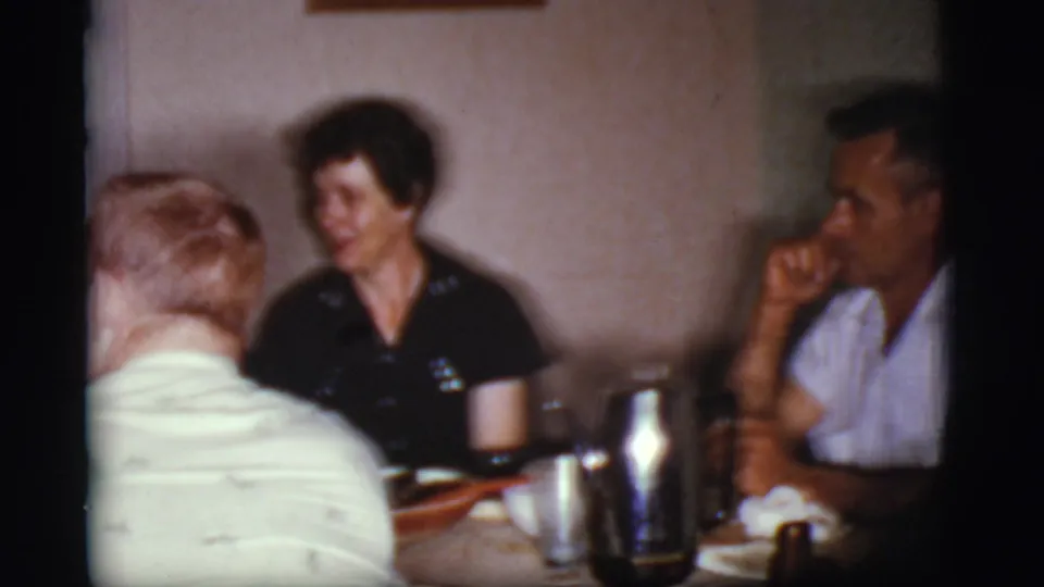 1968 Cottonwood, Arizona frame showing three adults around a dining table, with tableware, a coffee pot, and wall decor visible.