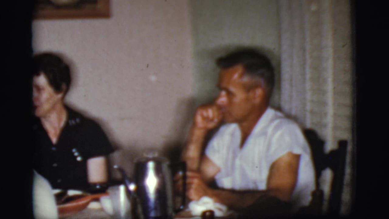 Two adults seated at a dining table with cups, dishes, and a metal pitcher in a dim room.