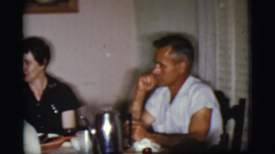 1968 Cottonwood, Arizona home-movie frame showing adults seated after a meal at a dining table with tableware and window blinds nearby.
