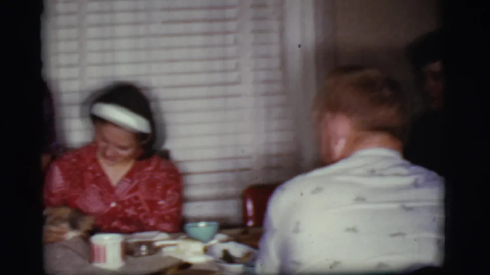 1968 Cottonwood, Arizona home-movie frame with a seated woman in a red patterned dress and white headband at a dining table.