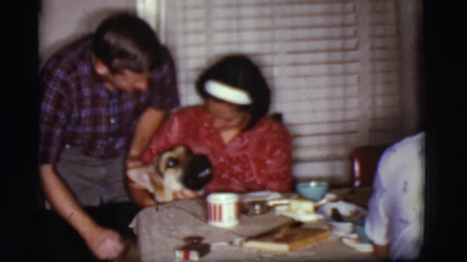 1968 Cottonwood, Arizona home-movie frame showing a man leaning in as a woman holds a dog near the dining table.