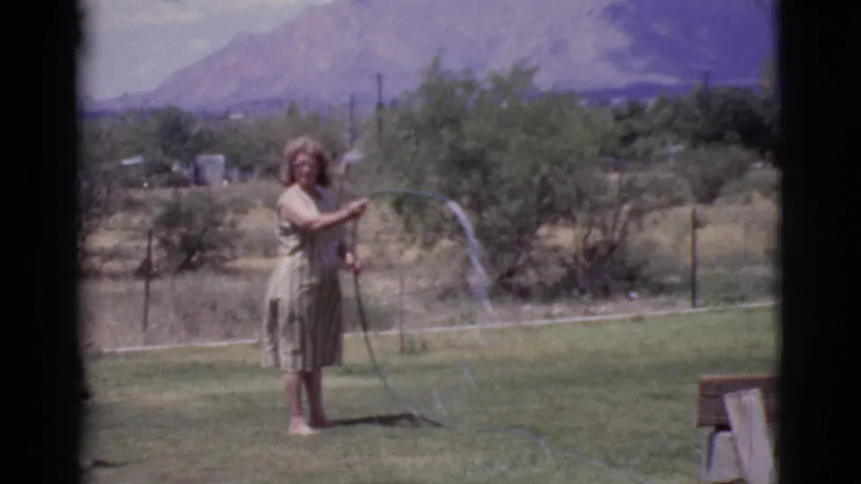 In a 1968 Cottonwood backyard, the woman points a hose across the grass with mountains and desert vegetation in the distance.