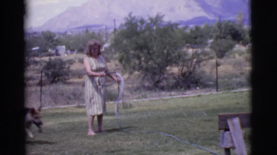 The woman stands barefoot in the Cottonwood yard with the hose looped in her hands as the German Shepherd moves near the edge of frame.