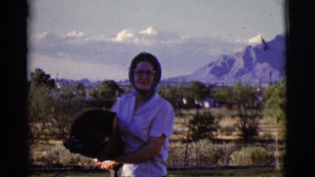Woman in a headscarf holds a dark hat outdoors with mountains behind her.