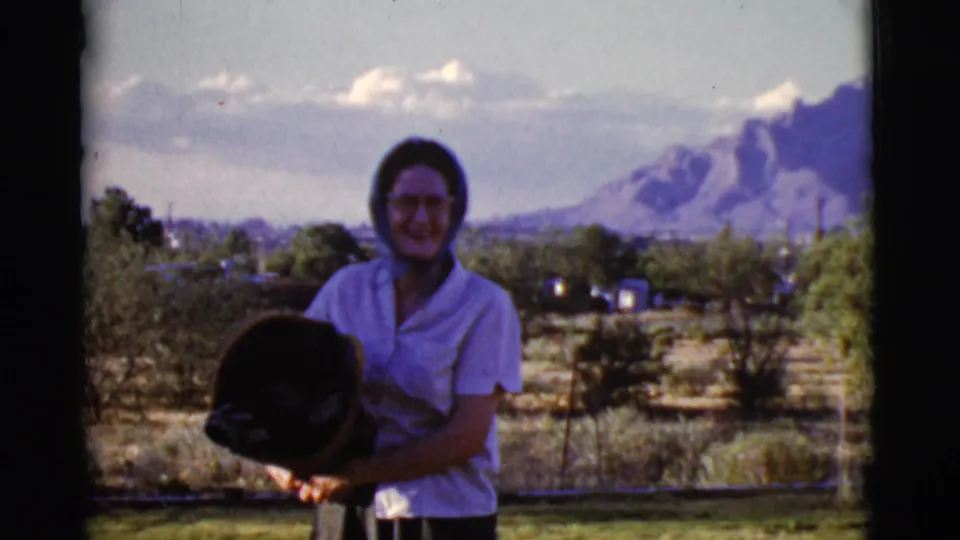 Smiling adult woman in a light blouse and headscarf stands outdoors in 1968 Cottonwood, Arizona, with mountains in the distance.