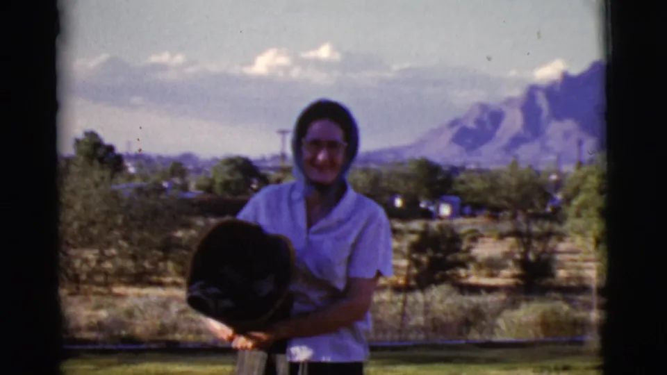 Adult woman in 1968 Cottonwood, Arizona, holds a dark hat and faces the camera against a faded 8mm mountain backdrop.