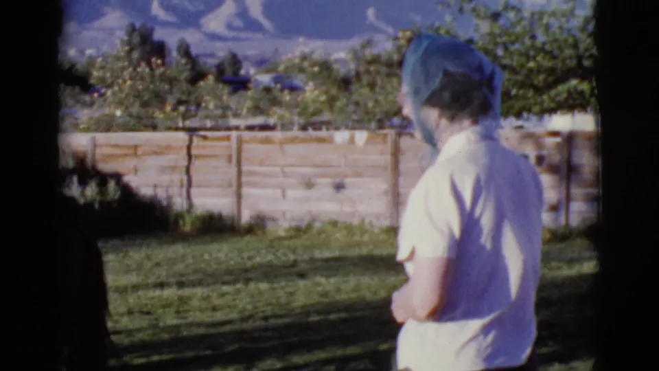 Woman in a white shirt and blue head covering faces away in a 1968 Cottonwood, Arizona backyard, with a block wall and mountains beyond.