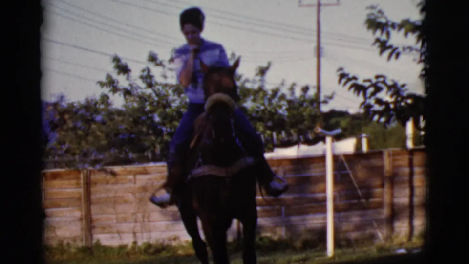 In Cottonwood, Arizona, a woman rides a dark horse near a wooden fence, with trees and utility lines visible in the 1968 home-movie frame.