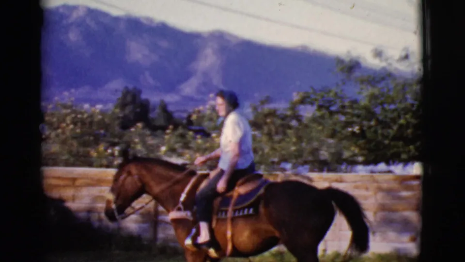 A 1968 Cottonwood, Arizona frame shows a woman riding a dark horse past a wooden fence, with blue mountains and trees in the background.